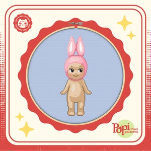 May include: A pixel art cross stitch pattern of a doll wearing a pink bunny hat. The pattern is in a red and white frame with a white background. The pattern is from Pop Pixel Patterns.
