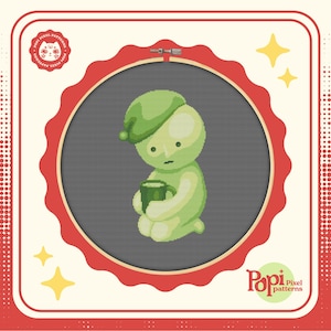 May include: A green pixel art character with a green hat and a cup in its hands. The character is sitting on a gray background inside a red embroidery hoop. The image is surrounded by a white border with a red and white polka dot pattern.