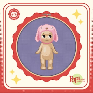 May include: A cross-stitch pattern featuring a cartoon baby wearing a pink bunny hat. The pattern is displayed in a red embroidery hoop on a purple background. The image includes the text "Pop! Pixel patterns".