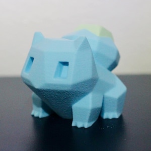 Könnte beinhalten: Eine geometrisch geformte Figur aus hellblauem Kunststoff, die einen Pokemon-Charakter darstellt. Die Figur hat einen abgerundeten, dreieckigen Kopf mit zwei kleinen, schwarzen Augen. Die Figur steht auf einer dunklen Oberfläche.