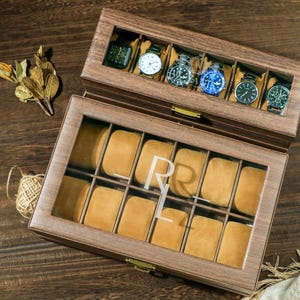 May include: A wooden watch box with a glass top and a bottom drawer. The top drawer has 6 slots for watches and the bottom drawer has 10 slots. The box is engraved with the initials "R.L."