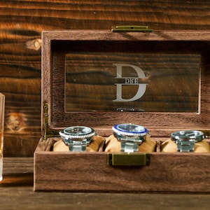 May include: A wooden watch box with a glass top and four compartments for storing watches. The box has a personalized inscription that reads "DBE".