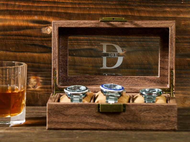May include: A wooden watch box with a glass top and four compartments for storing watches. The box has a personalized inscription that reads "DBE".