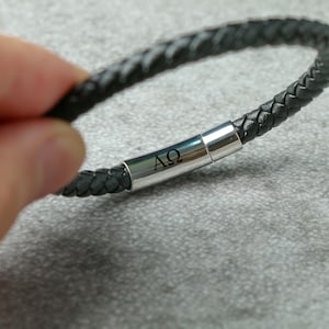 Könnte beinhalten: Schwarzes Lederarmband mit einem silbernen Verschluss, der mit den Buchstaben "AΩ" graviert ist.