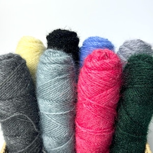 May include: A wicker basket filled with eight skeins of yarn in various colours, including black, yellow, light blue, grey, pink, and green.