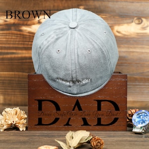 Puede incluir: Una caja de madera marrón con la palabra "DAD" grabada en la parte delantera. La caja tiene una ranura para una gorra de béisbol, y una gorra de béisbol gris se muestra en la ranura. El texto "Drive Safe, We Love You Dad!" está grabado debajo de la palabra "DAD".