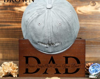 Puede incluir: Una caja de madera marrón con la palabra "DAD" grabada en la parte delantera. La caja tiene una ranura para una gorra de béisbol, y una gorra de béisbol gris se muestra en la ranura. El texto "Drive Safe, We Love You Dad!" está grabado debajo de la palabra "DAD".