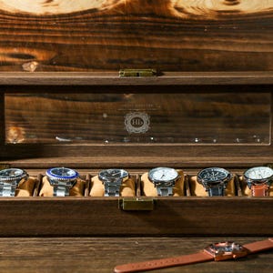 May include: A wooden watch box with a glass top and eight slots for watches. The box is closed and the watches are visible through the glass. The box is made of dark wood and has a brass latch.