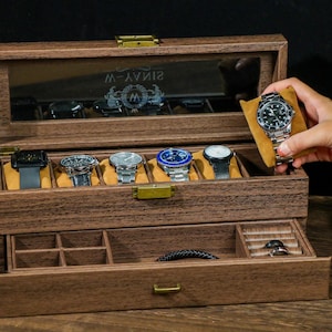 May include: A wooden watch box with a glass top and a drawer. The box has a brown finish and is engraved with the text "SINAVY-W". The box has six slots for watches and a drawer for storing other accessories.