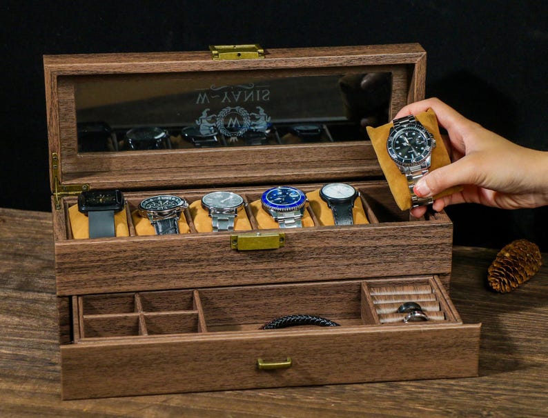 May include: A wooden watch box with a glass top and a drawer. The box has a brown finish and is engraved with the text "SINAVY-W". The box has six slots for watches and a drawer for storing other accessories.