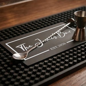 Può includere: Un allestimento bar con un tappetino nero con superficie strutturata. Un cucchiaio da bar in acciaio inossidabile, un doppio jigger e un cartello nero con la scritta "The James Bar EST. 2026" sono sul tappetino. Lo sfondo è una superficie in legno.
