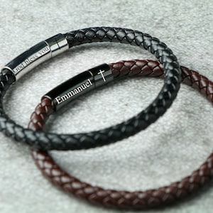 Könnte beinhalten: Zwei Lederarmbänder mit Metallakzenten. Ein Armband ist schwarz und sagt "Gott segne dich" und das andere ist braun und sagt "Emmanuel" mit einem Kreuz.