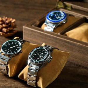 May include: Three silver wristwatches with black faces and silver bands are displayed in a wooden watch box. The box has a brown velvet lining and is open to show the watches.