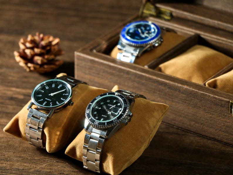 May include: Three silver wristwatches with black faces and silver bands are displayed in a wooden watch box. The box has a brown velvet lining and is open to show the watches.