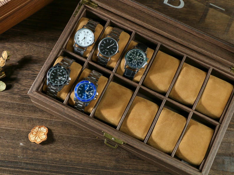May include: A wooden watch box with a clear lid, featuring 10 compartments for storing watches. The box is made of dark brown wood and has a velvet lining. There are four watches in the box, with different styles and colors.
