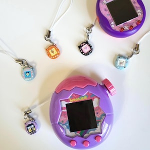 May include: A collection of vintage Tamagotchi virtual pet devices in purple, pink, orange, black, and blue. The devices have small screens and attached keychains. The larger device has the text "Tamagotchi" on the front.