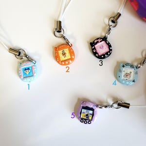 May include: A collection of five colorful, miniature handheld game console charms. Each charm features a small screen with a cartoon graphic and is attached to a white lanyard with a metal clasp. The charms are in various colors, including blue, orange, black, and purple.