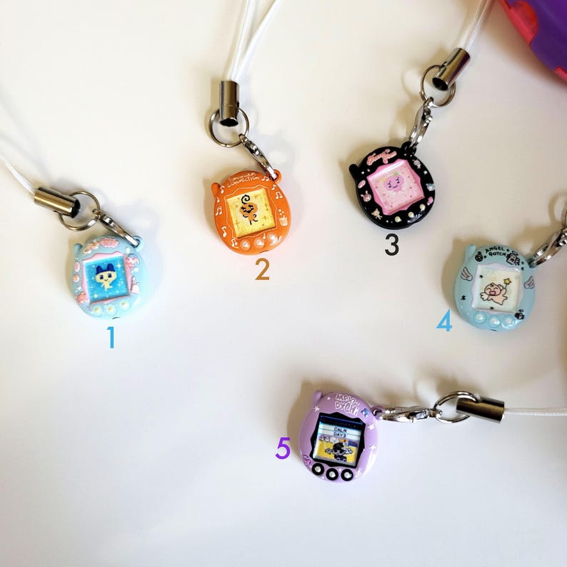 May include: A collection of five colorful, miniature handheld game console charms. Each charm features a small screen with a cartoon graphic and is attached to a white lanyard with a metal clasp. The charms are in various colors, including blue, orange, black, and purple.