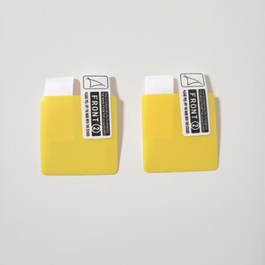 May include: Two yellow screen protectors with white tabs. Each protector has a label that reads "FRONT 2" and instructions to peel off the mask with the sticker. The protectors are designed to safeguard a device's screen.