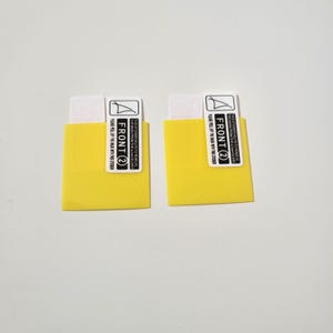 May include: Two yellow screen protector applicators with white and black labels. The labels read "FRONT 2" and include instructions to peel off the mask with the sticker. The applicators are designed to protect screens.