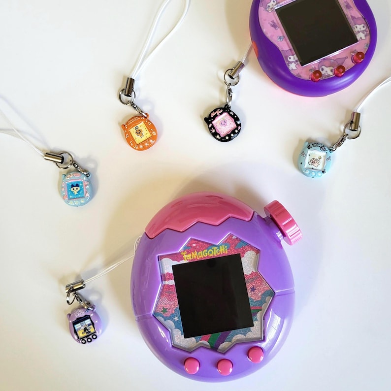 May include: A collection of vintage Tamagotchi virtual pet devices in purple, pink, orange, black, and blue. The devices have small screens and attached keychains. The larger device has the text "Tamagotchi" on the front.
