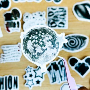 May include: A silver disco ball sticker with a white border and silver glitter stars. The sticker is on a wooden surface with other stickers.