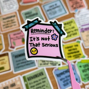 May include: A pink reminder note sticker with a black border and a green and blue plaid design. The note says "Reminder: It's not that serious" and has a smiley face and a purple flower.