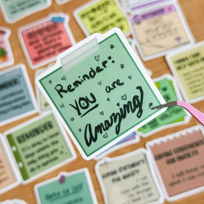 May include: Green sticker with a white border and rounded corners. The sticker has a handwritten message that says "Reminder: You are Amazing" with hearts and stars around the text.
