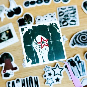 May include: A black and white sticker with a woman's face and a red star over her eye. The sticker is on a wooden surface with other stickers.