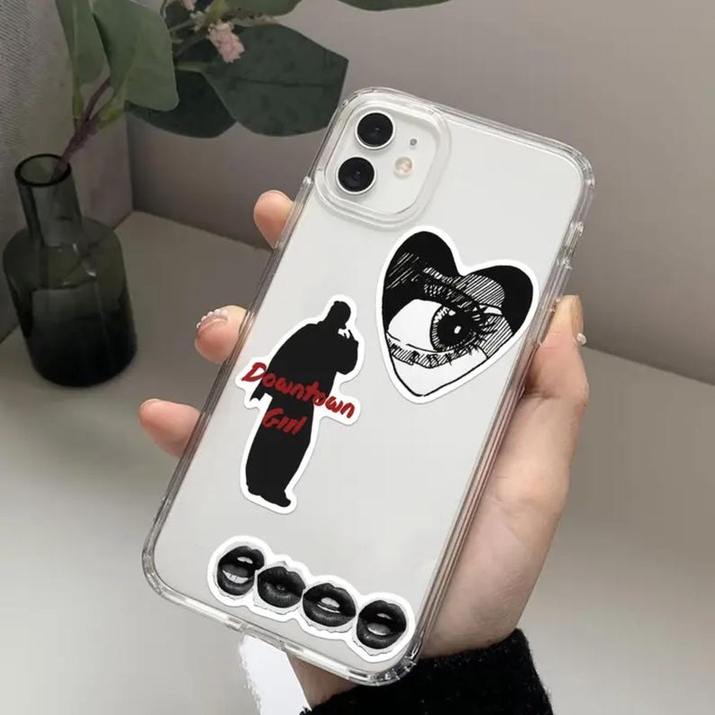 May include: A clear phone case with black and white stickers. The stickers include a heart with an eye, a silhouette of a person with the text "Downtown Girl", and three lips.