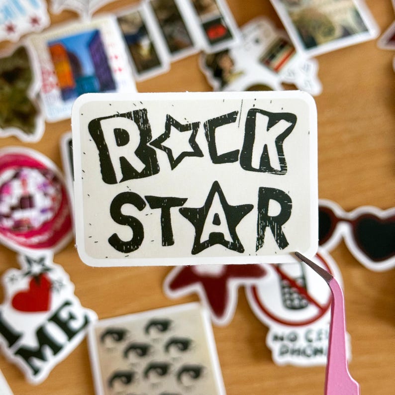 以下が含まれることがあります： 白地に黒文字で「Rock Star」と書かれたステッカー。星が単語の間にあります。ステッカーは使い古したような見た目で、少しすり切れています。