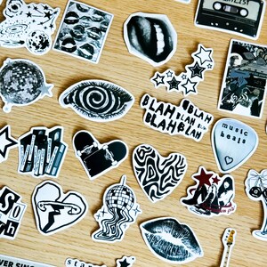 May include: A collection of black and white stickers featuring various designs, including a spiral eye, a heart with wavy lines, a skateboard, a disco ball, a music note, and lips. The stickers are arranged on a wooden surface.