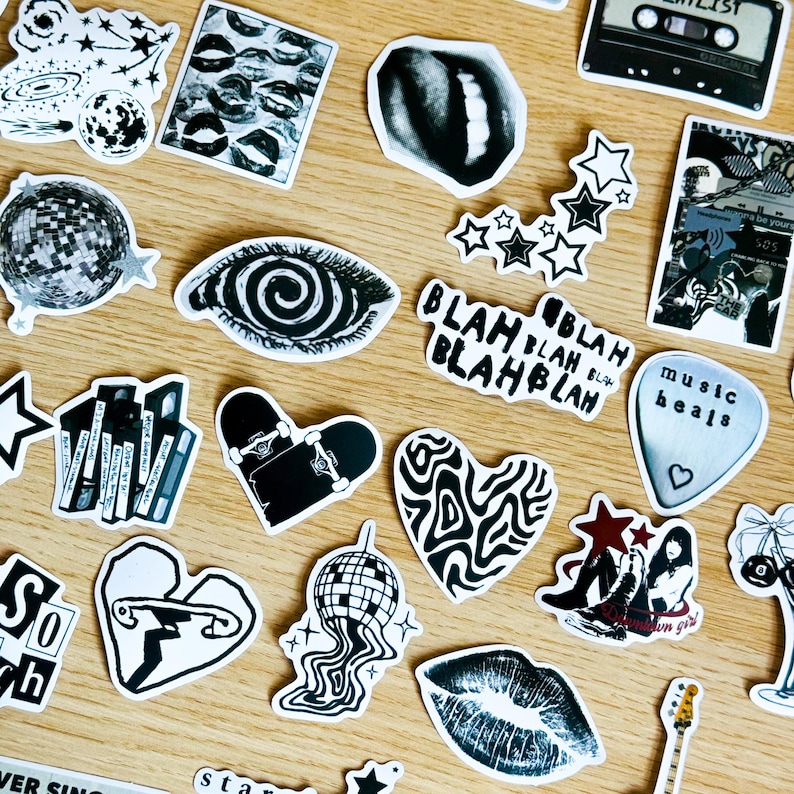 May include: A collection of black and white stickers featuring various designs, including a spiral eye, a heart with wavy lines, a skateboard, a disco ball, a music note, and lips. The stickers are arranged on a wooden surface.