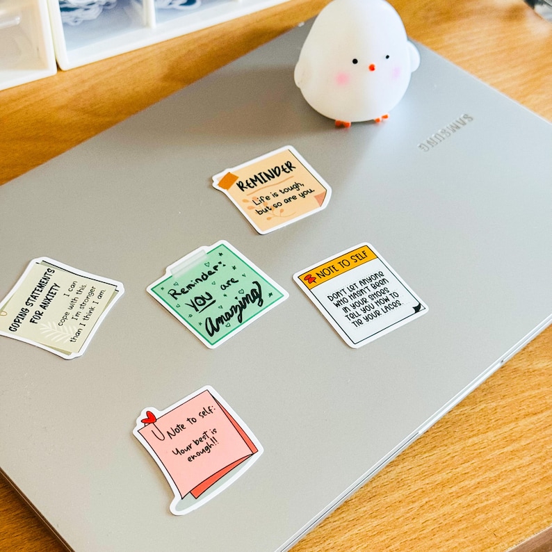 May include: Five colourful sticky notes with motivational messages on a silver laptop. The notes say "Reminder: You are Amazing", "Reminder: Life is tough, but so are you", "Note to self: Your best is enough", "Coping Statements for Anxiety: I can cope with this. I'm stronger than I think I am", and "Note to self: Don't let anyone who hasn't been in your shoes tell you how to tie your laces".