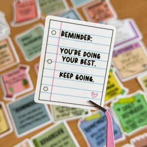 May include: A white sticker with pink lines and a pink heart. The sticker has the text "REMINDER: YOU'RE DOING YOUR BEST. KEEP GOING."