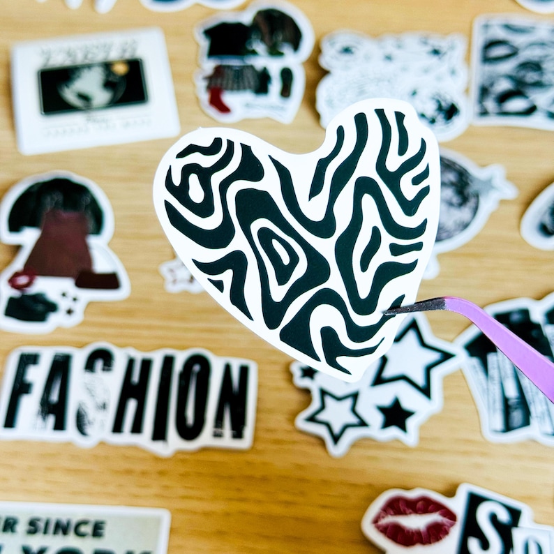 May include: A black and white heart-shaped sticker with a swirling pattern. The sticker is being held by tweezers.