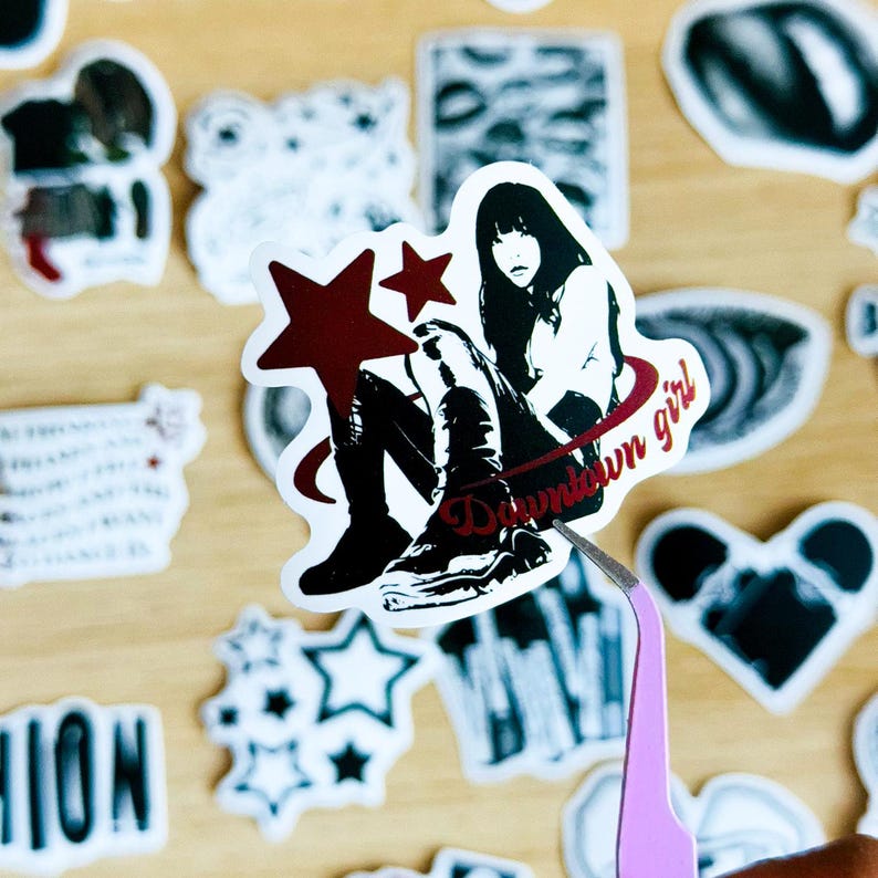 May include: A sticker featuring a cartoon girl with black hair and a red star. The girl is wearing a black and white outfit and is sitting on a red circle with the text "Downtown girl" written on it.