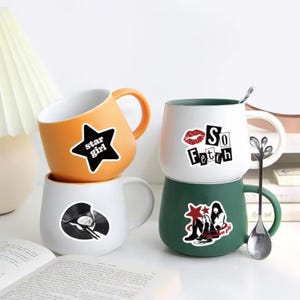 May include: Three ceramic mugs with different designs. The first mug is orange with a black star and the text "star girl". The second mug is white with a red lip and the text "So Fetch". The third mug is green with a black and white illustration of a woman sitting with the text "On fleek girl".
