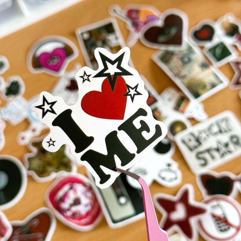 以下が含まれることがあります： 赤いハート、黒い文字で「I LOVE ME」と書いてある黒い星がついた白いステッカー。ステッカーは他のステッカーのあるテーブルの上に置かれています。