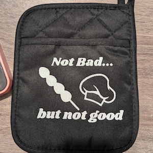 May include: Black quilted oven mitt with a white design that says "Not Bad... but not good" with a chef's hat and a skewer of food.