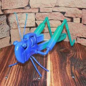 May include: A blue and green metal grasshopper sculpture made from recycled materials. The grasshopper is posed with its legs outstretched and its antennae pointing upwards.