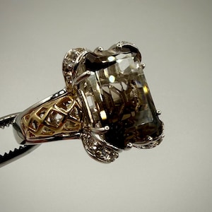 May include: A large, rectangular-cut smoky quartz gemstone ring. The ring features a silver band with gold filigree accents and small, clear accent stones. The gemstone is held in place by a silver setting with decorative details.