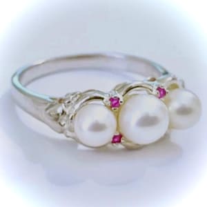 May include: A silver ring featuring three large, white pearls. Small, square, red gemstones are set between the pearls. The ring has a detailed, ornate design on the sides, and is set against a white background.