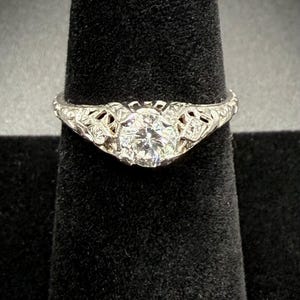May include: A silver engagement ring with a large, round, clear gemstone. The ring features intricate filigree detailing around the center stone and along the band. The ring is displayed on a black velvet surface.