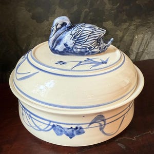 May include: A white ceramic lidded serving dish with blue painted accents. The lid is topped with a blue and white swan figurine. The dish has a round shape with a decorative blue floral pattern around the rim and base.