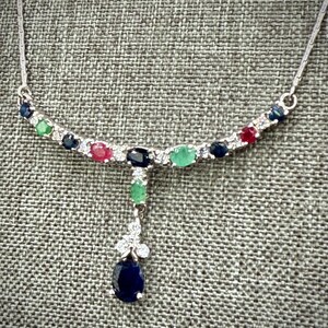 May include: A silver necklace with a curved bar design, adorned with a series of colorful gemstones. The gemstones include blue, green, and red stones, along with clear accent stones. A teardrop-shaped, dark blue gemstone hangs from the center.