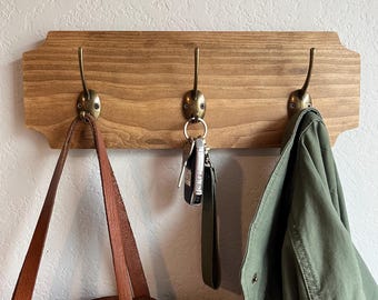 May include: A wooden wall-mounted coat rack with three hooks. A brown leather tote bag hangs on one hook, a set of keys hangs on another, and a green jacket hangs on the third hook.