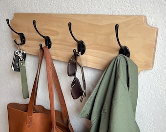May include: A wooden wall-mounted coat rack with four black metal hooks. A brown leather tote bag, sunglasses, and a green jacket are hanging on the hooks.
