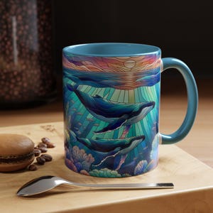 May include: A blue ceramic mug with a colorful stained glass design of whales swimming in the ocean. The design features a sunset with a sun shining through the water.