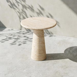 May include: A round side table crafted from beige travertine stone, featuring a smooth circular tabletop supported by a solid, tapered cylindrical base. The natural porous texture of the stone is visible across the entire surface of the piece.